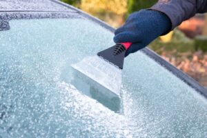 A gloved hand uses a small scraper to remove ice from a car windshield